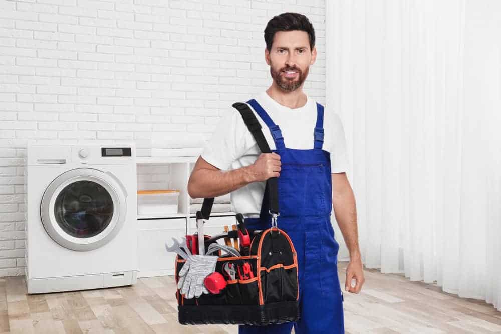 A man in blue overalls and a white shirt stands indoors holding a tool bag. He has dark hair and a beard. Behind him is a washing machine, white brick wall, and sheer curtains.