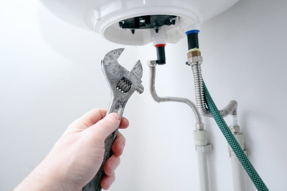 A hand holding an adjustable wrench near pipes connected to the underside of a water heater, preparing to perform maintenance or repair.