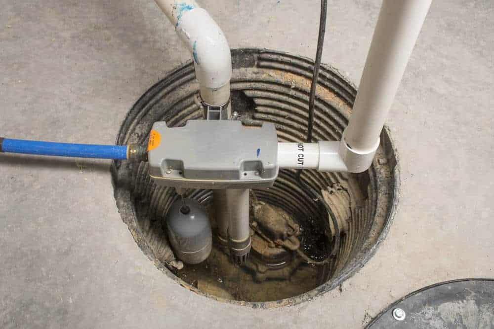 A close-up view of a sump pump system installed in a basement floor pit, with connected PVC pipes for water inlet and outlet, and electrical components visible above the pump.