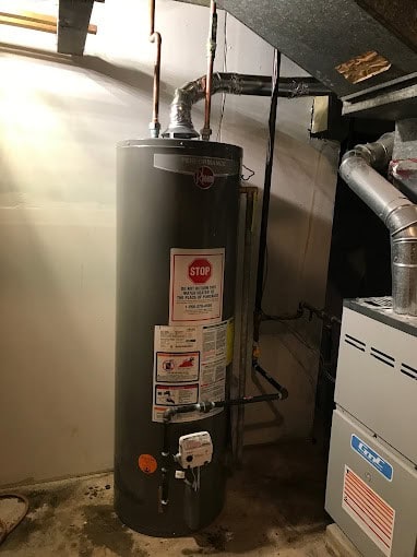 A gray water heater with warning labels is installed in a basement corner near a furnace, with exposed pipes and ductwork visible on the wall and ceiling.